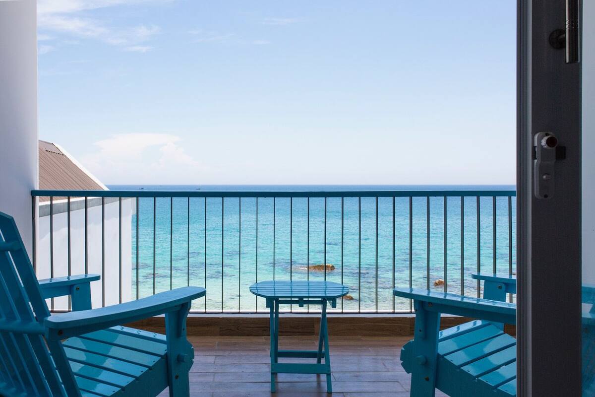 A balcony with two blue Adirondack chairs and a small table, overlooking a clear blue sea.