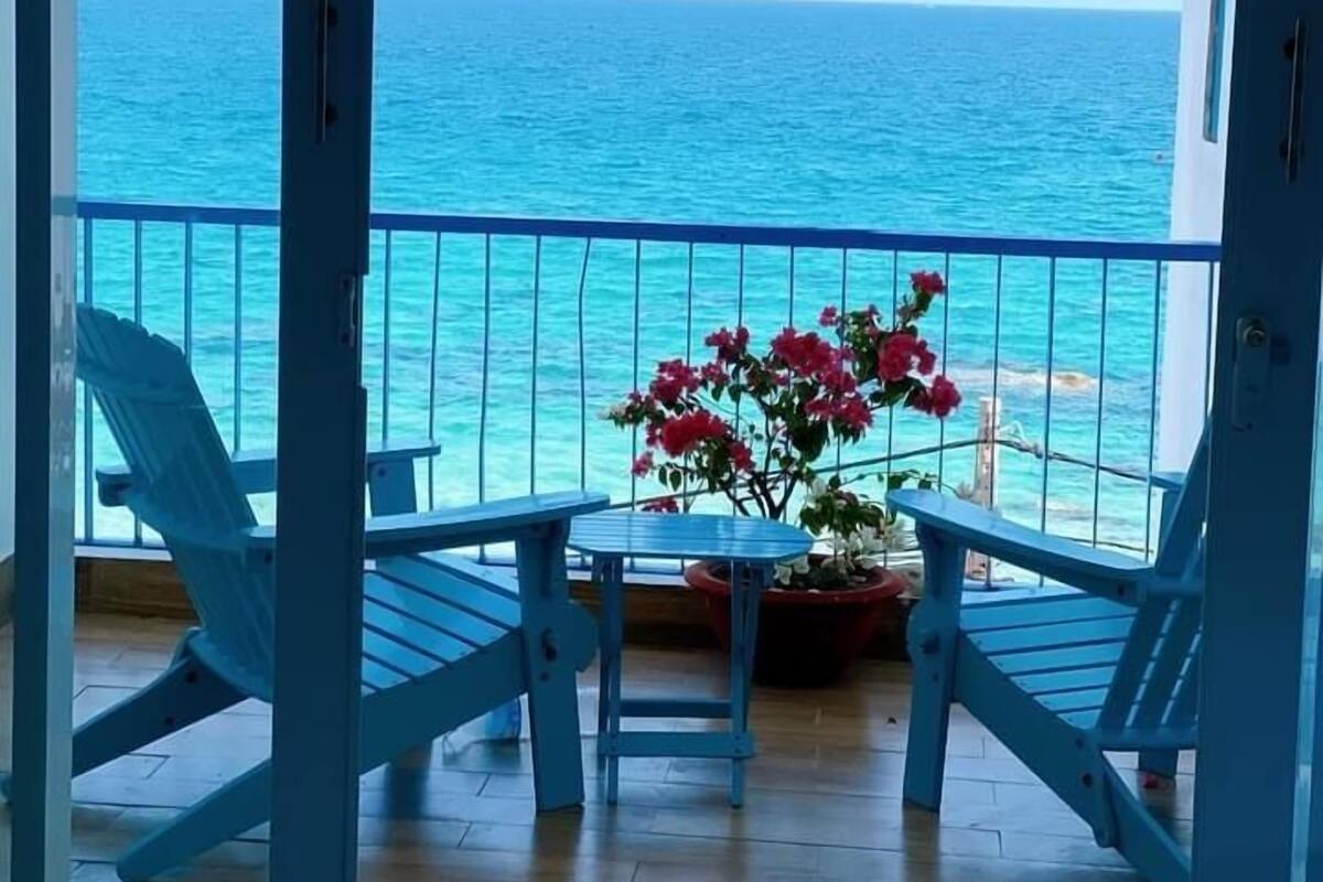 A balcony with a view of the ocean, featuring two blue chairs, a small table, and a potted plant with pink flowers.