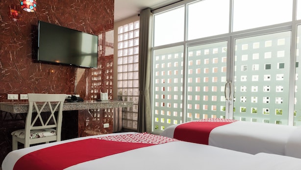 Habitación de hotel con dos camas, un escritorio, una silla, un televisor y ventanas grandes con vistas a un edificio.