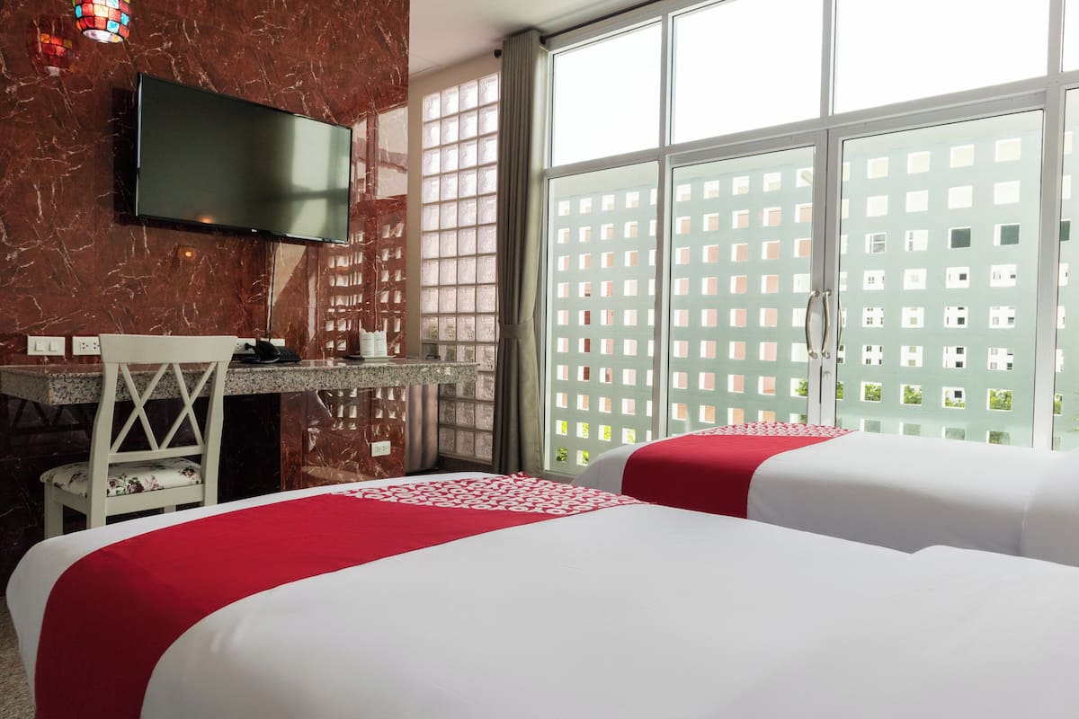 A hotel room with two beds, a desk, a chair, a TV, and large windows with a view of a building.