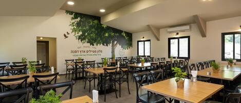 A spacious restaurant with multiple wooden tables and black chairs, featuring a large mural and potted plants.