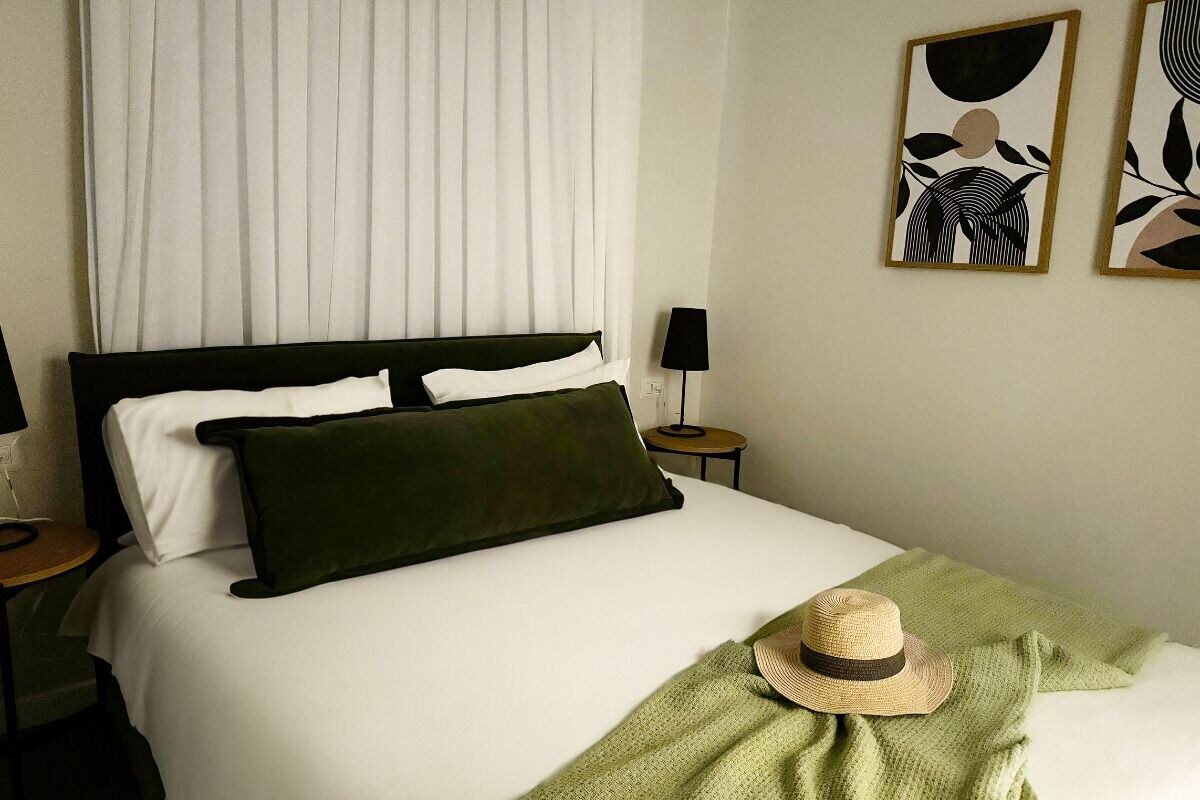 Una cama bien hecha con almohadas blancas y verde oscuro, una manta verde y un sombrero de paja sobre la cama. Hay dos obras de arte enmarcadas en la pared.