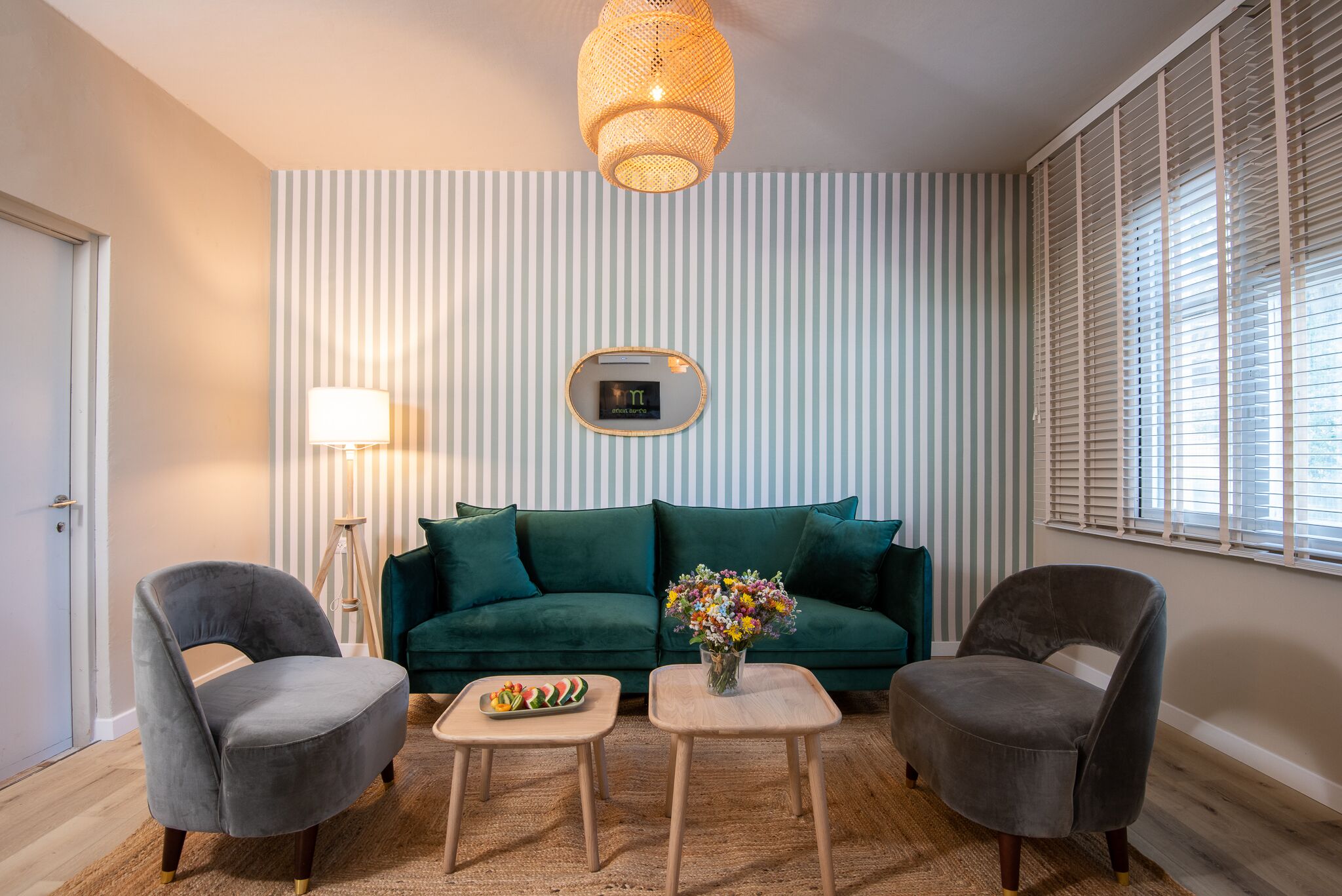 A living room with a teal sofa, two grey armchairs, a small wooden coffee table, a floor lamp, a wall-mounted thermostat, and a window with blinds.