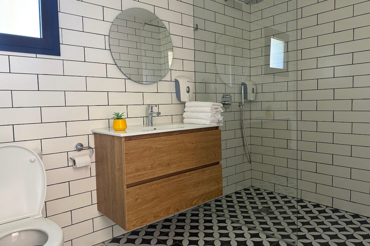 A bathroom with white tiled walls, a wooden vanity, a toilet, a sink, and a shower.
