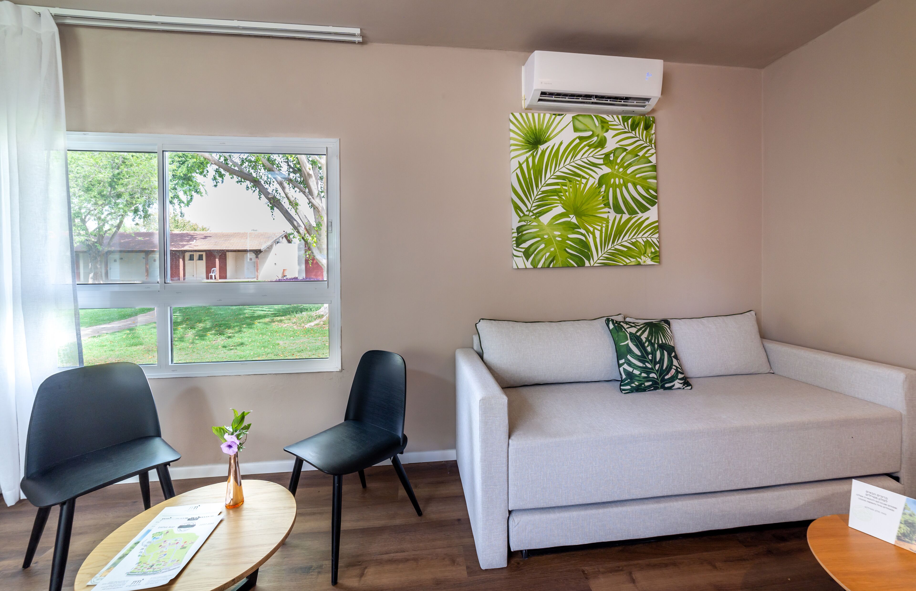 Een kamer met een bank, twee stoelen, een kleintje tafeltje met een vaas, een raam met uitzicht op een gebouw en bomen, en een airconditioning.