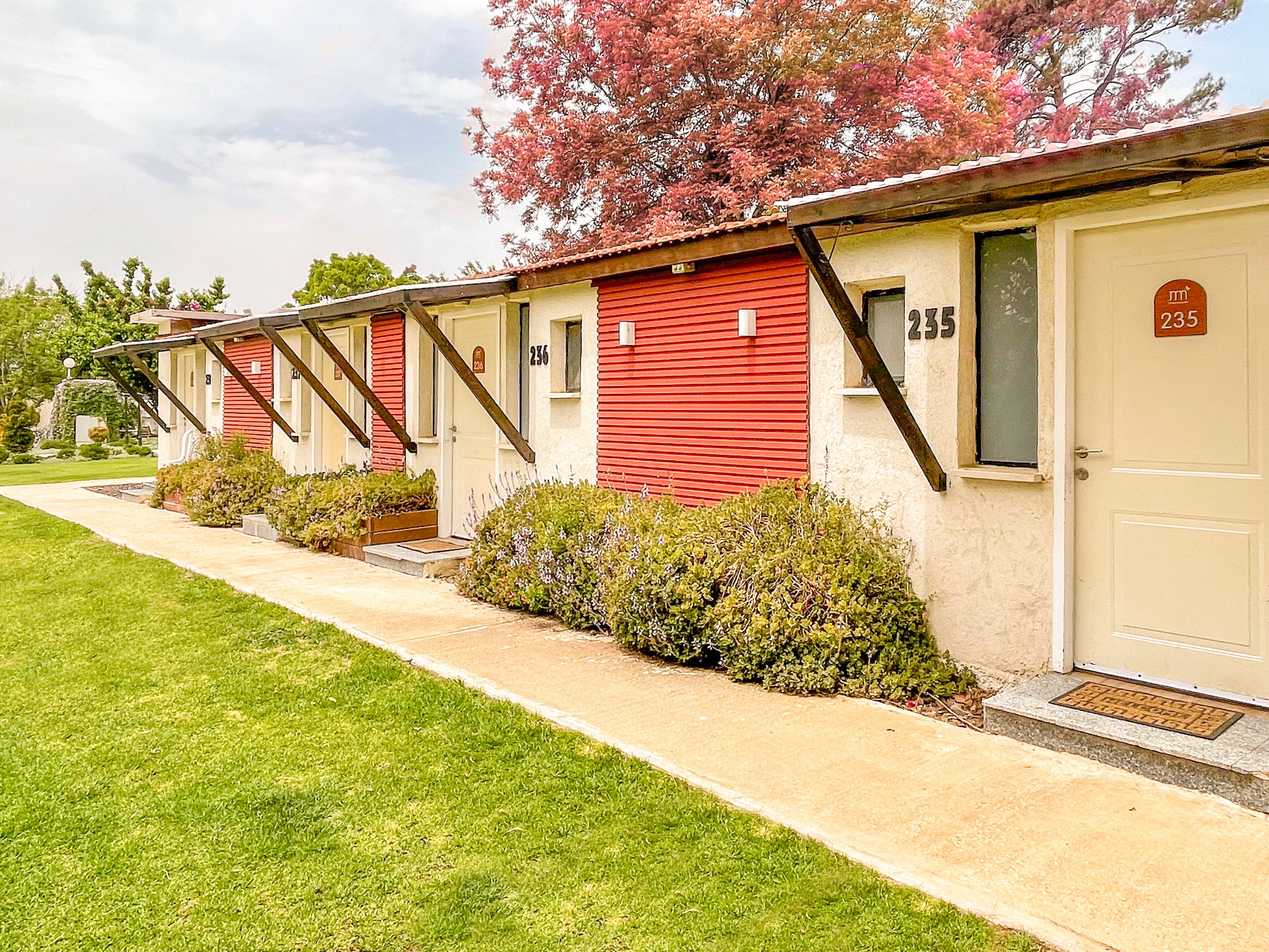 A row of motel rooms with red exteriors and beige doors, numbered 235 and 236, featuring a simple, functional design with small front yards and a clear sky.
