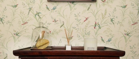Una consola de madera con una jaula de pájaro de vidrio, un difusor de caña y una vela con la etiqueta 'aroma botánico' frente a un papel tapiz con motivos de pájaros y plantas.