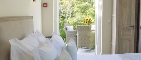 A bedroom with a large bed, a nightstand, a wall-mounted lamp, and an open door leading to a balcony with outdoor furniture and sunflowers.