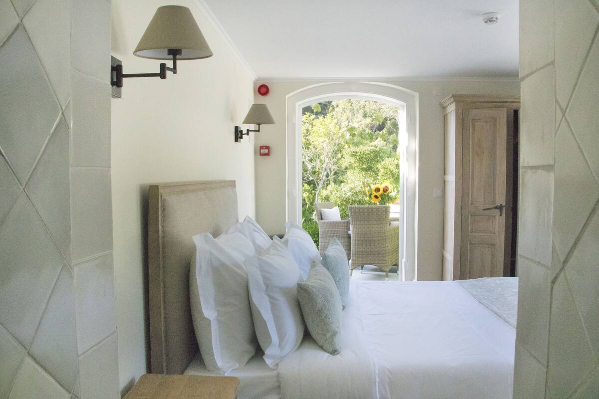 Un dormitorio con cama, almohadas, una lámpara de pared y vistas a la vegetación.