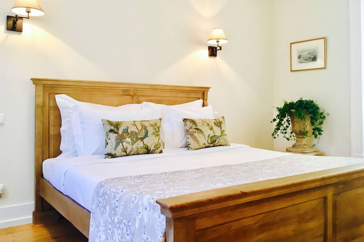 Una cama bien hecha con sábanas blancas y dos almohadones decorativos, un cabecero de madera y dos lámparas de pared.