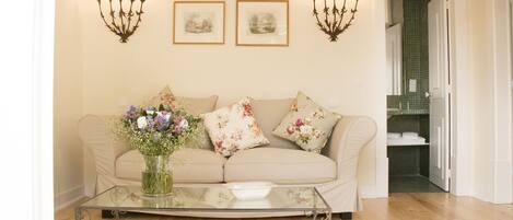 A living room with a beige sofa, floral cushions, a glass coffee table with books, a vase with flowers, and wall-mounted candelabras.