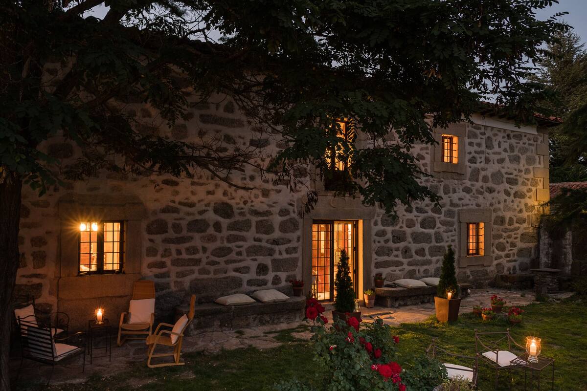 Un edificio de piedra con jardín, mesas al aire libre y un interior iluminado.