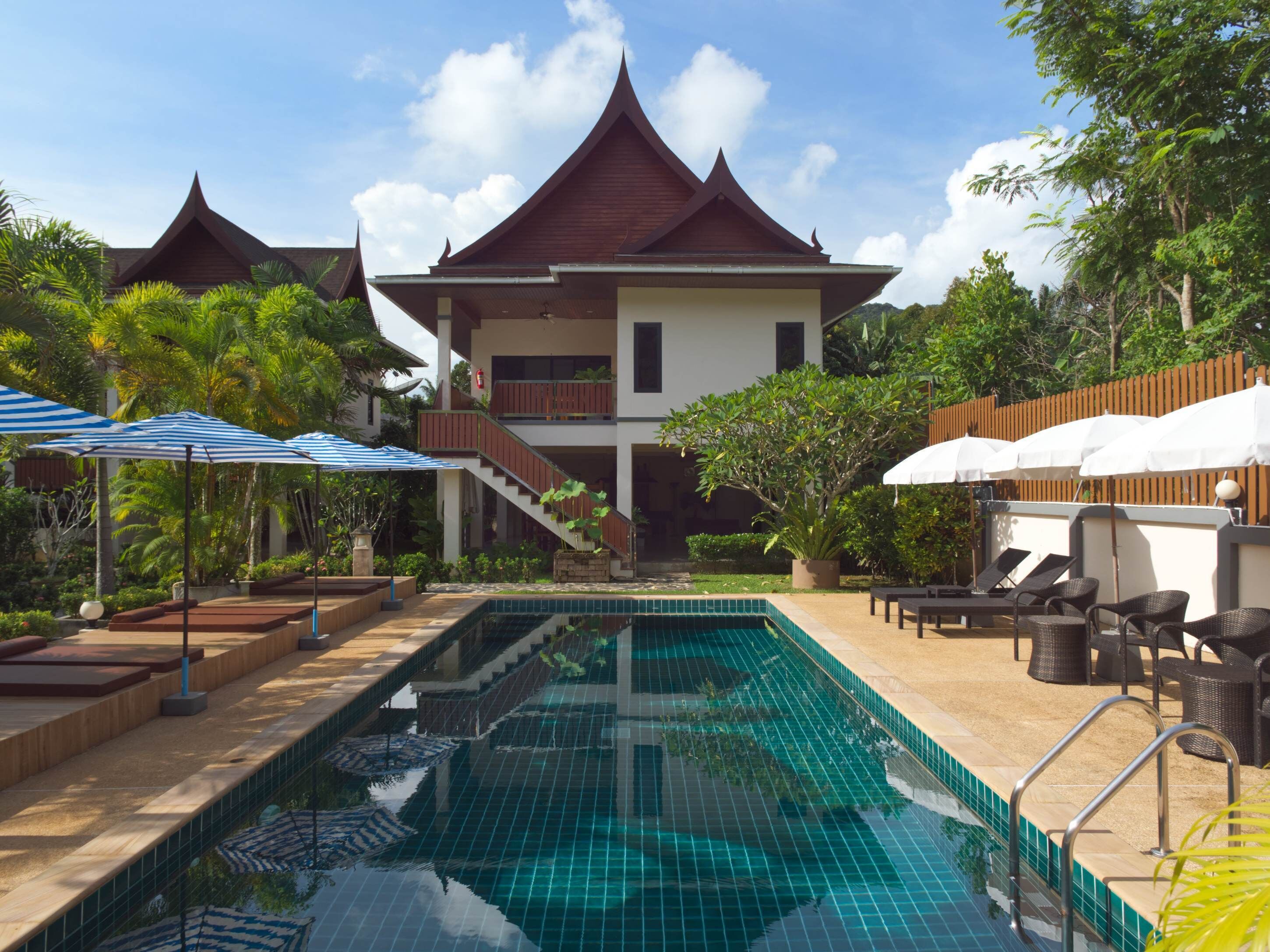 Piscina com espreguiçadeiras e guarda-sóis, rodeada por vegetação exuberante e um edifício tradicional com telhado inclinado.