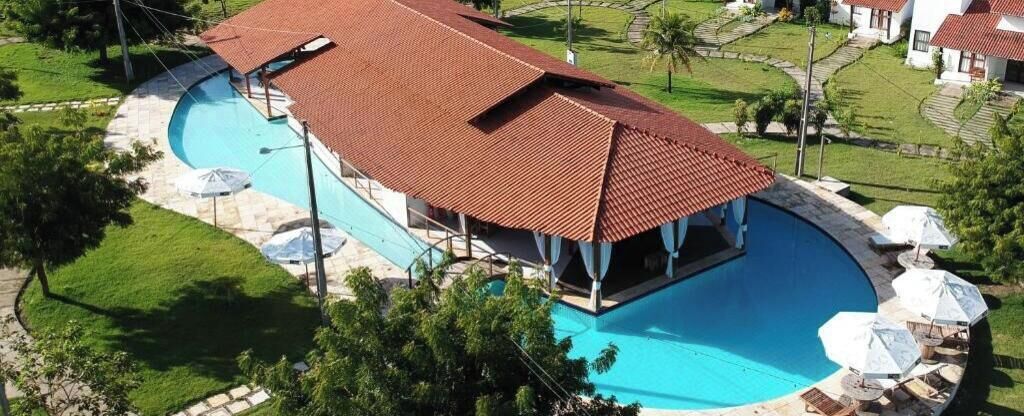 Piscina com área coberta, cercada por um gramado bem cuidado e alguns guarda-sóis.