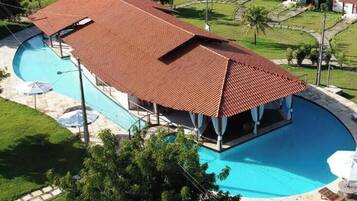 Piscina com área coberta, cercada por um gramado bem cuidado e alguns guarda-sóis.