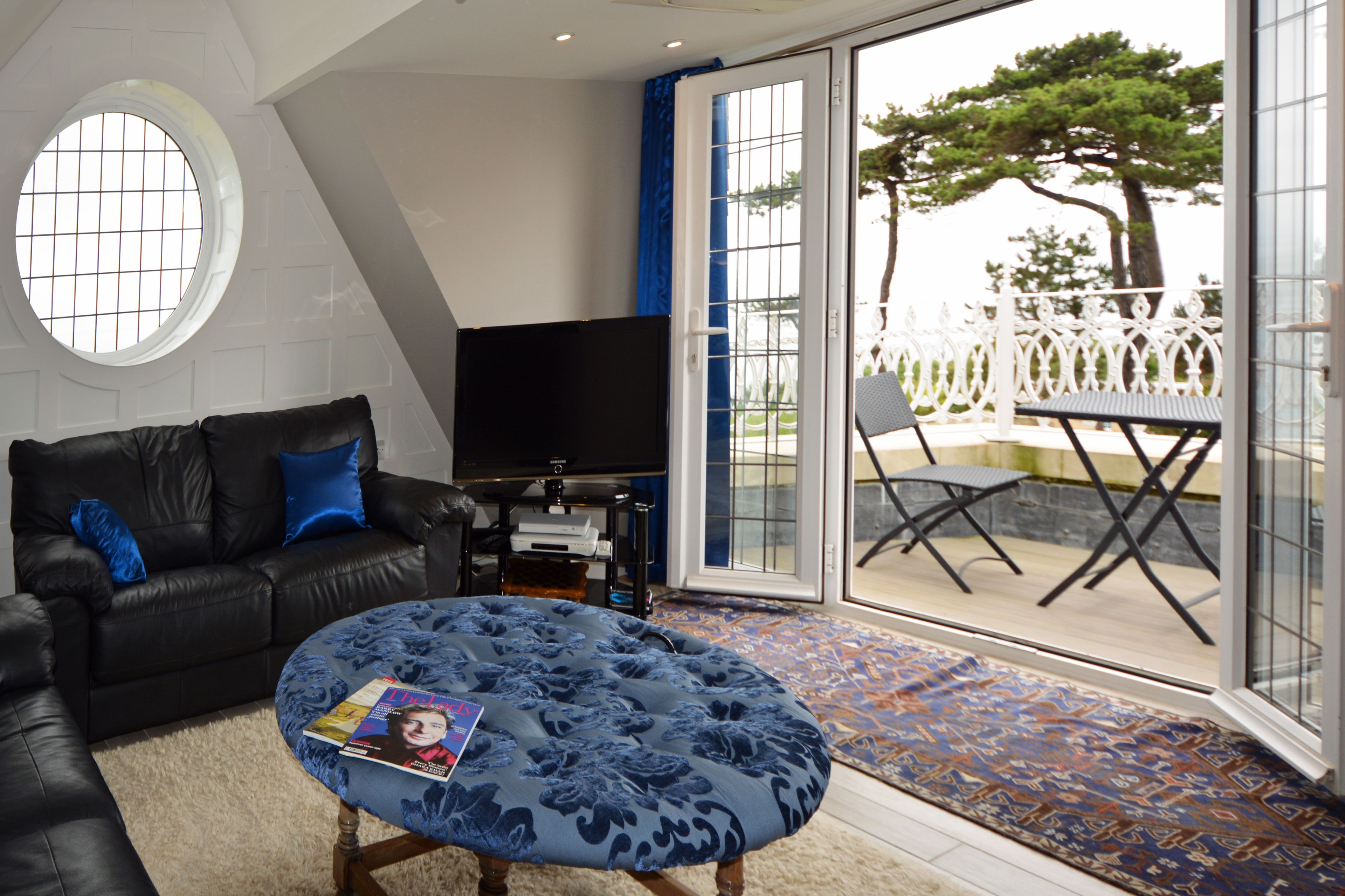 A living room with a black leather sofa, a round ottoman with a magazine, a flat-screen TV, and a large sliding glass door leading to an outdoor patio with a white fence and a tree.