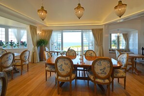 Un comedor con una mesa grande y sillas, ventanas amplias con cortinas y candelabros de cristal colgantes.