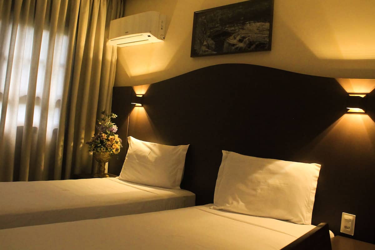A hotel room with two beds, a nightstand, a wall-mounted air conditioner, and a painting on the wall.