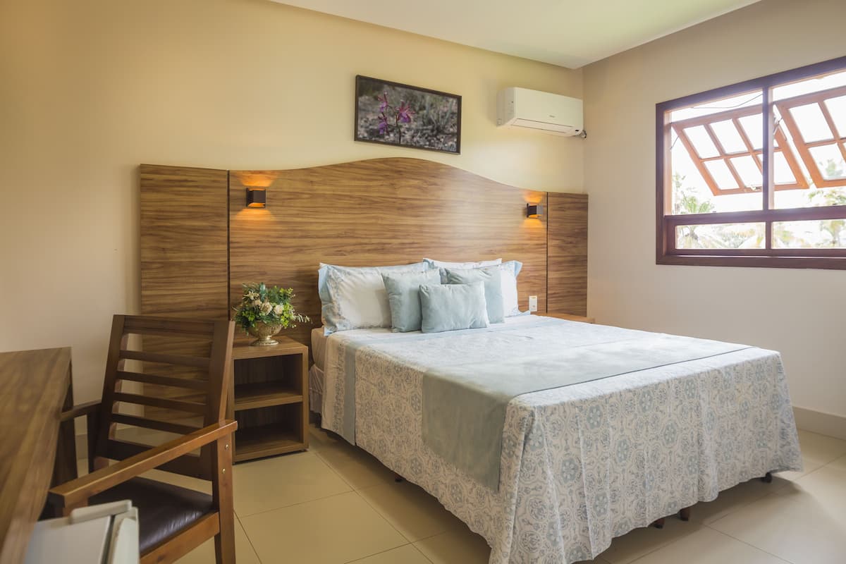 A bedroom with a wooden headboard, a bed with patterned bedding, a nightstand with a lamp, and a window with a view of greenery.
