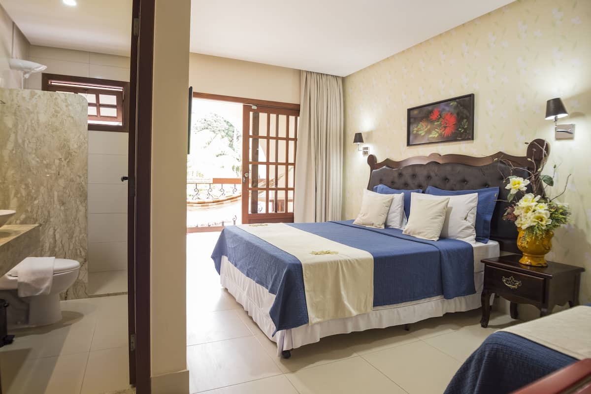 A bedroom with a bed, a nightstand, a bathroom with a toilet, and a view of the outside.