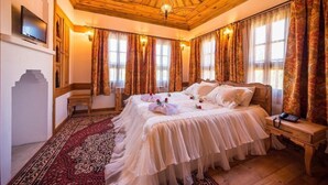 Ein traditionell eingerichtetes Zimmer mit Holzbalkendecke, einem großen Bett mit weißer Bettwäsche, einem roten Teppich mit Muster und Vorhängen mit Paisley-Design.