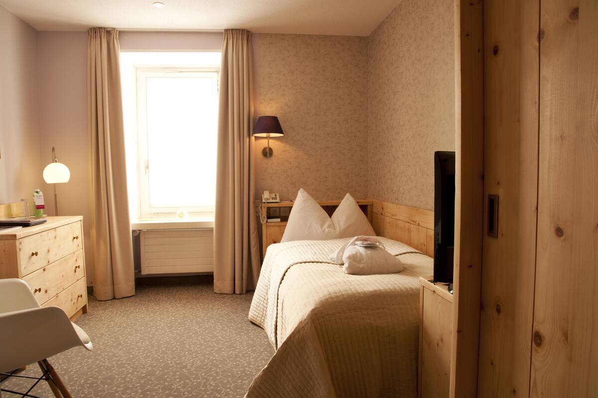 A hotel room with a bed, a desk with a lamp, a chair, and a window with curtains.