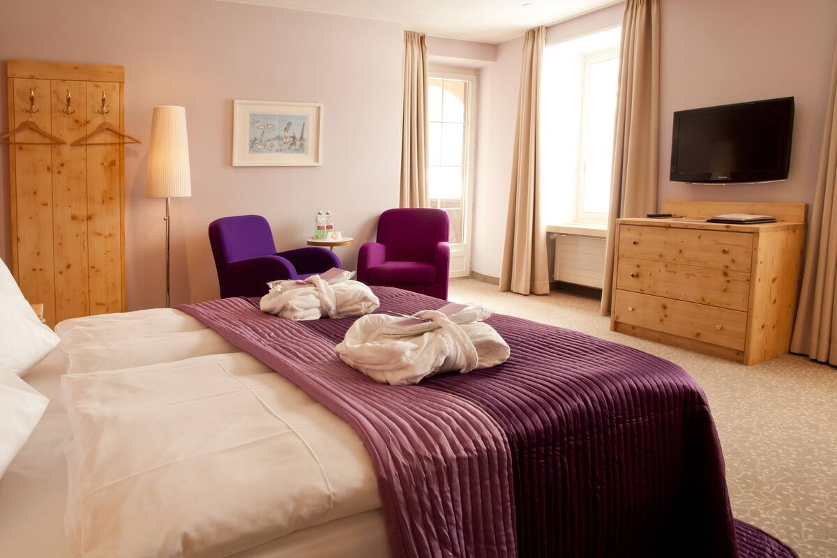 A hotel room with a bed, two chairs, a dresser, a television, and a window with curtains.