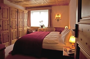 A hotel room with a large bed, a desk with a lamp, a chair, a small table with a plant, and wooden paneling on the walls.