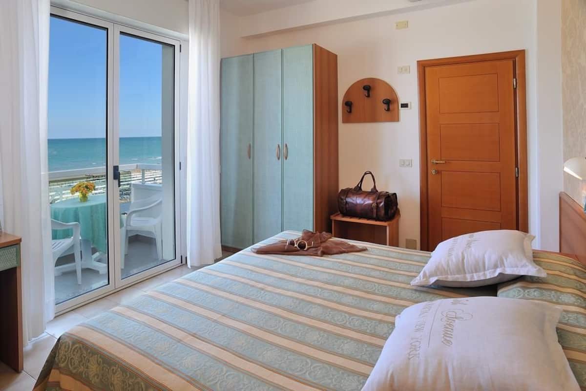 A hotel room with a bed, a wooden door, a coat rack, and a view of the beach through a sliding door.