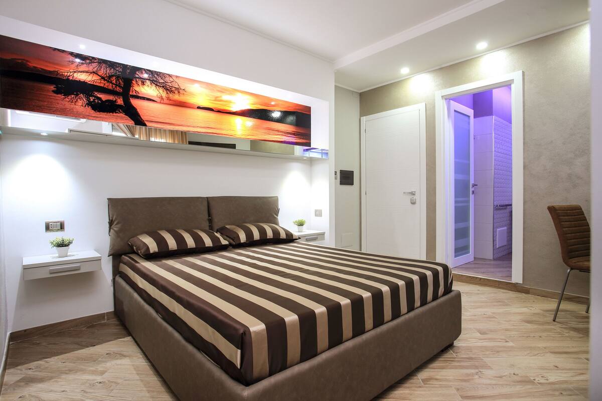 Un dormitorio moderno con una cama grande, una mesita de noche, un televisor de pared y una puerta que da a un baño.