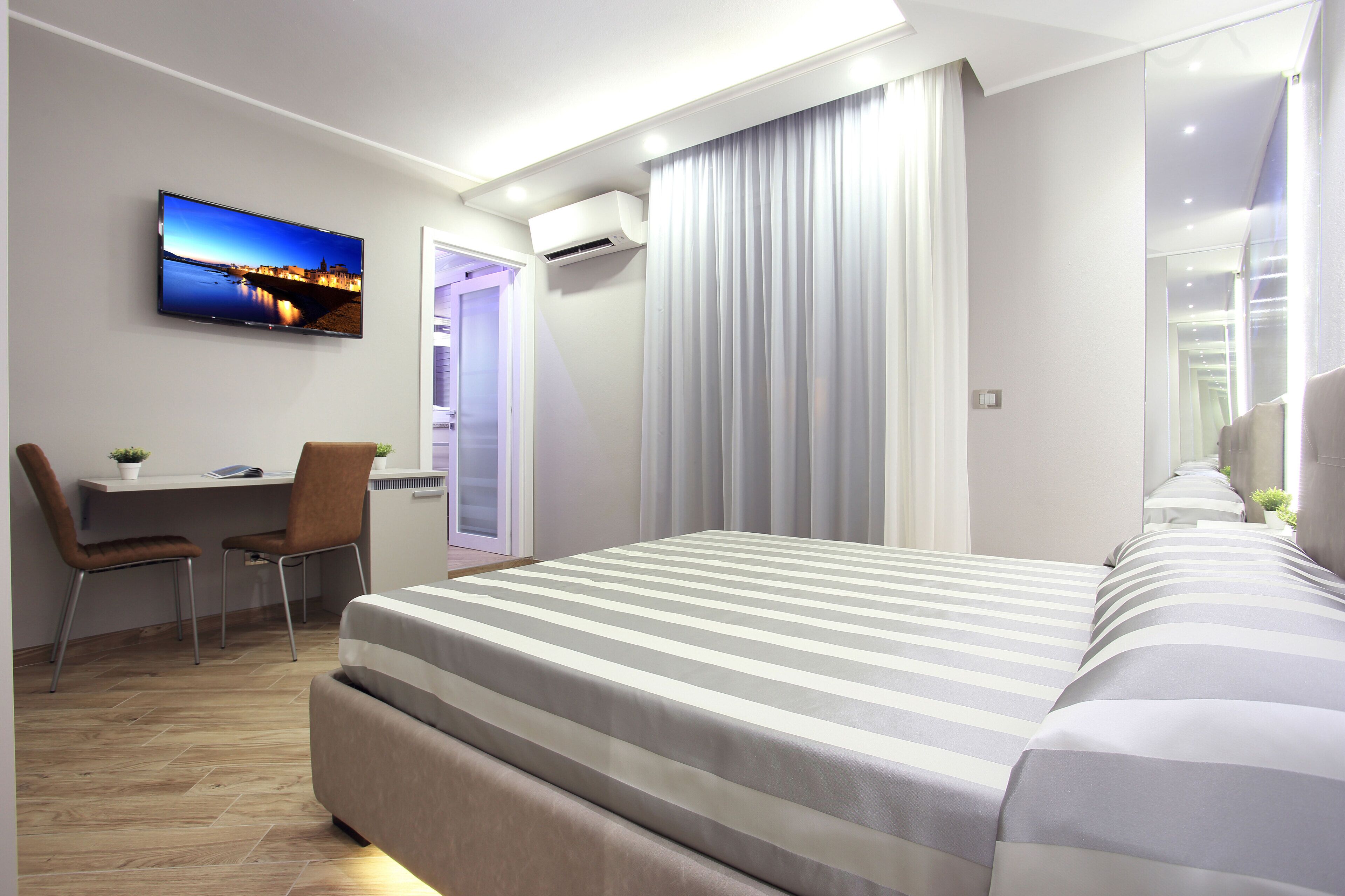A modern hotel room with a large bed, a desk with chairs, a TV on the wall, and an air conditioning unit.