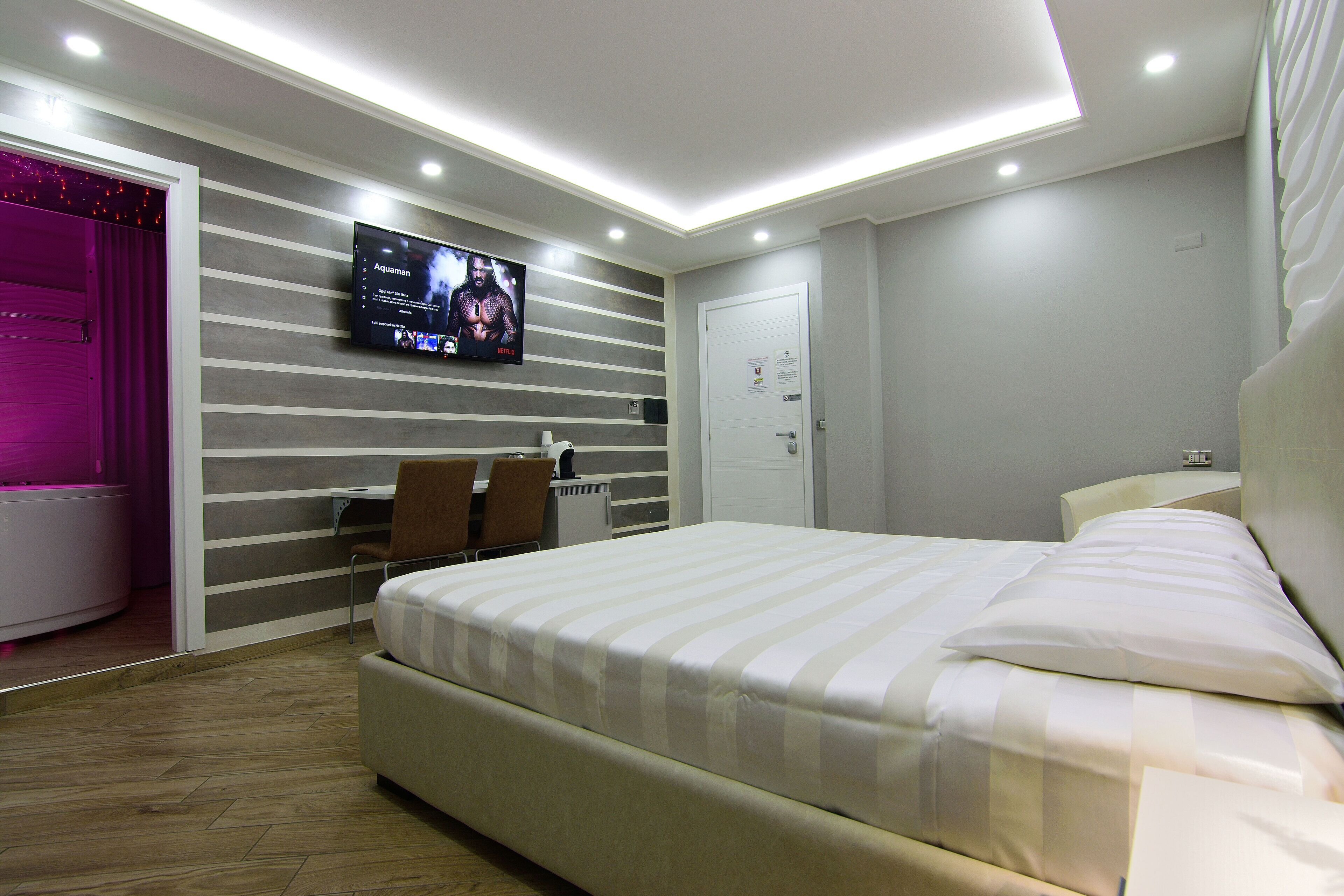 Una habitación de hotel moderna con una cama grande, un televisor de pantalla plana montado en la pared, un escritorio con una silla y un baño con zona de ducha iluminada en rosa.