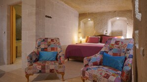 Une pièce avec deux fauteuils rembourrés, un lit recouvert d’une couverture rouge et un mur en pierre.