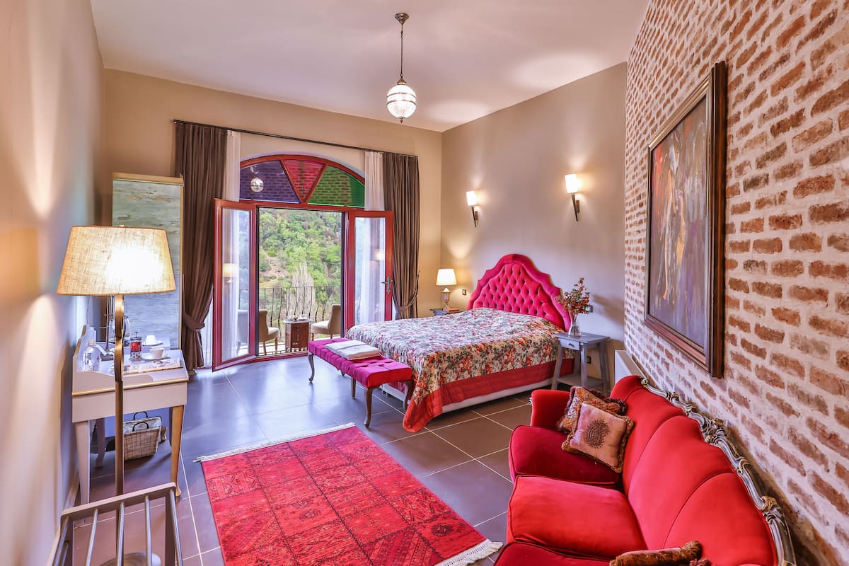 A bedroom with a bed, a red sofa, a desk with a lamp, a red rug, and a view of the outdoors through a glass door.