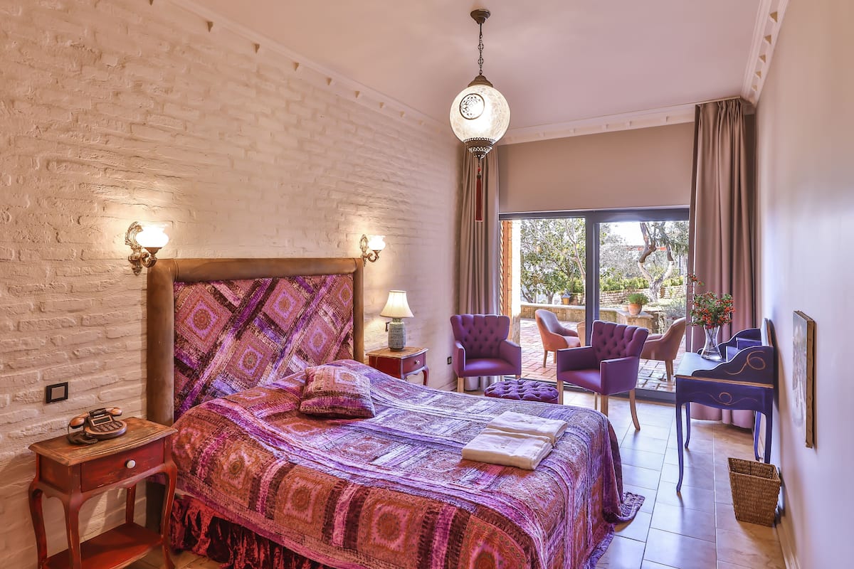 A bedroom with a large bed, a wooden nightstand, a lamp, and a view of the outside.