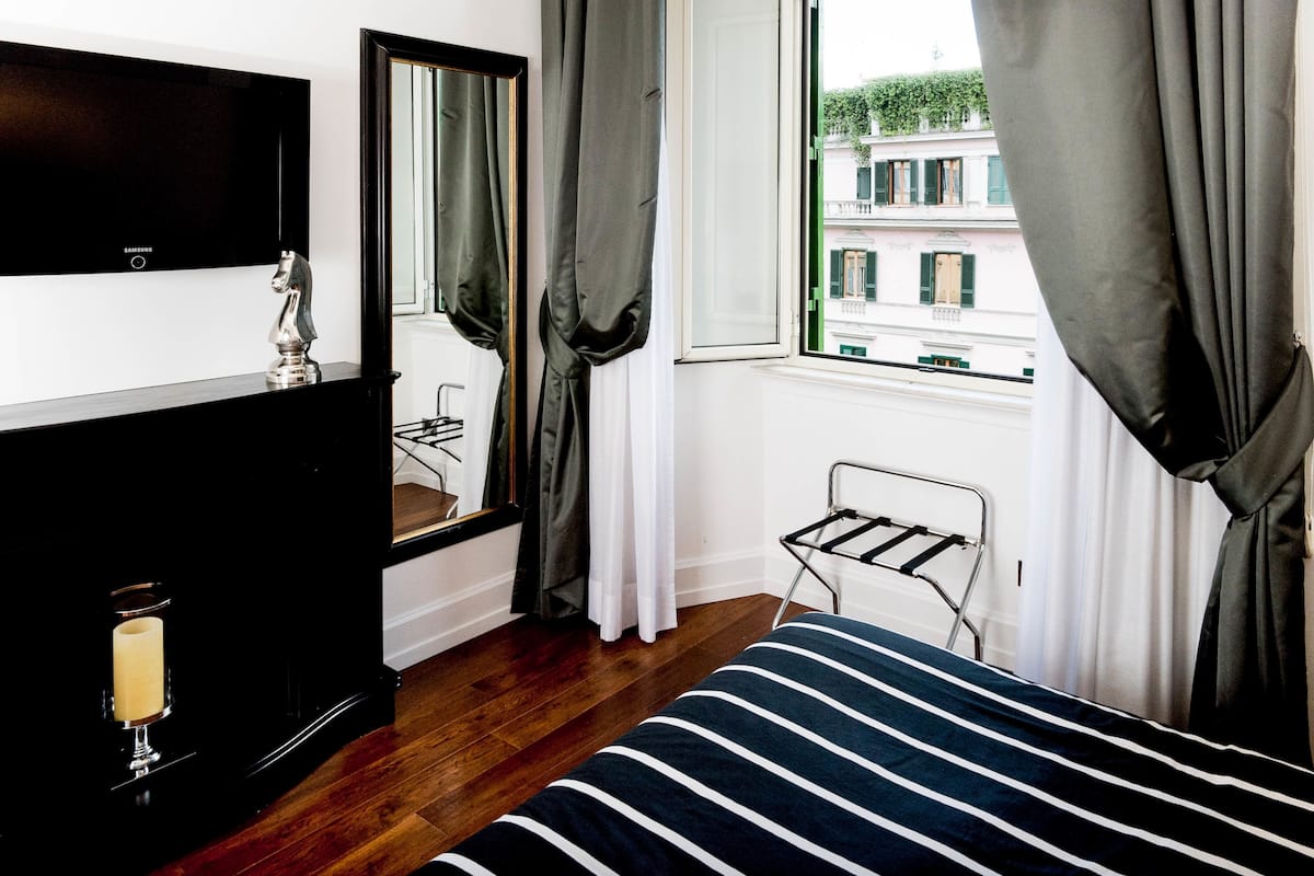 A hotel room with a bed, a TV, a mirror, a window with curtains, and a small metal rack.