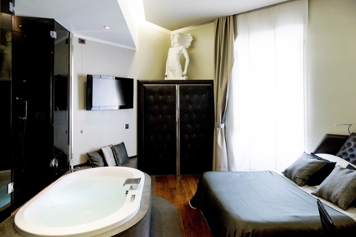 A modern hotel room with a large bed, a separate sitting area, a bathtub, and a statue on the wall.