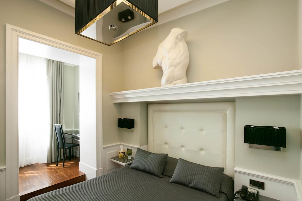 A bedroom with a large bed, a white headboard, a wall-mounted sculpture, a bedside table with a lamp, and a telephone.