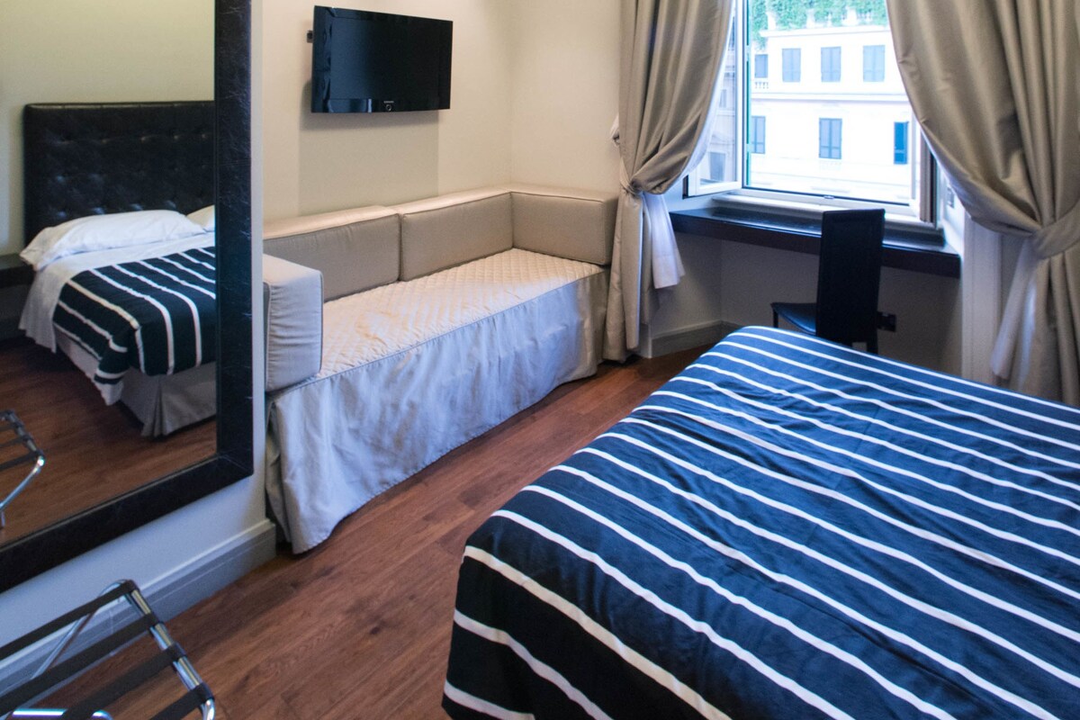 A hotel room with a bed, a desk, a chair, a TV mounted on the wall, and a window with curtains.