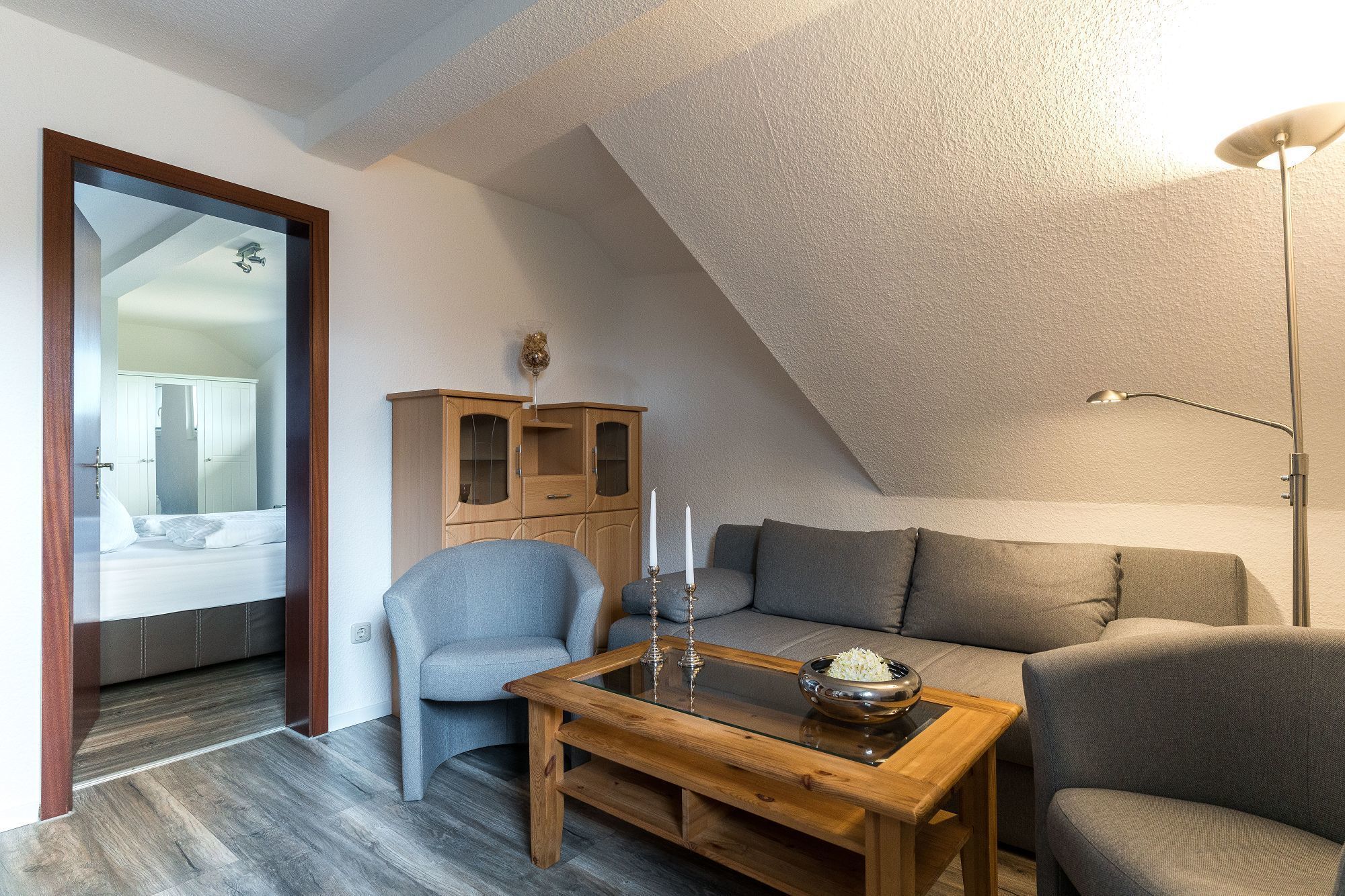 A cozy loft bedroom with a sloped ceiling, a wooden cabinet, a grey armchair, a grey sofa, and a glass-top coffee table with a bowl of rice.