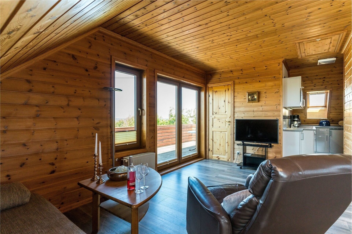 A cozy cabin interior with a wooden ceiling, a dining area with a table and chairs, a television, and a kitchen area.