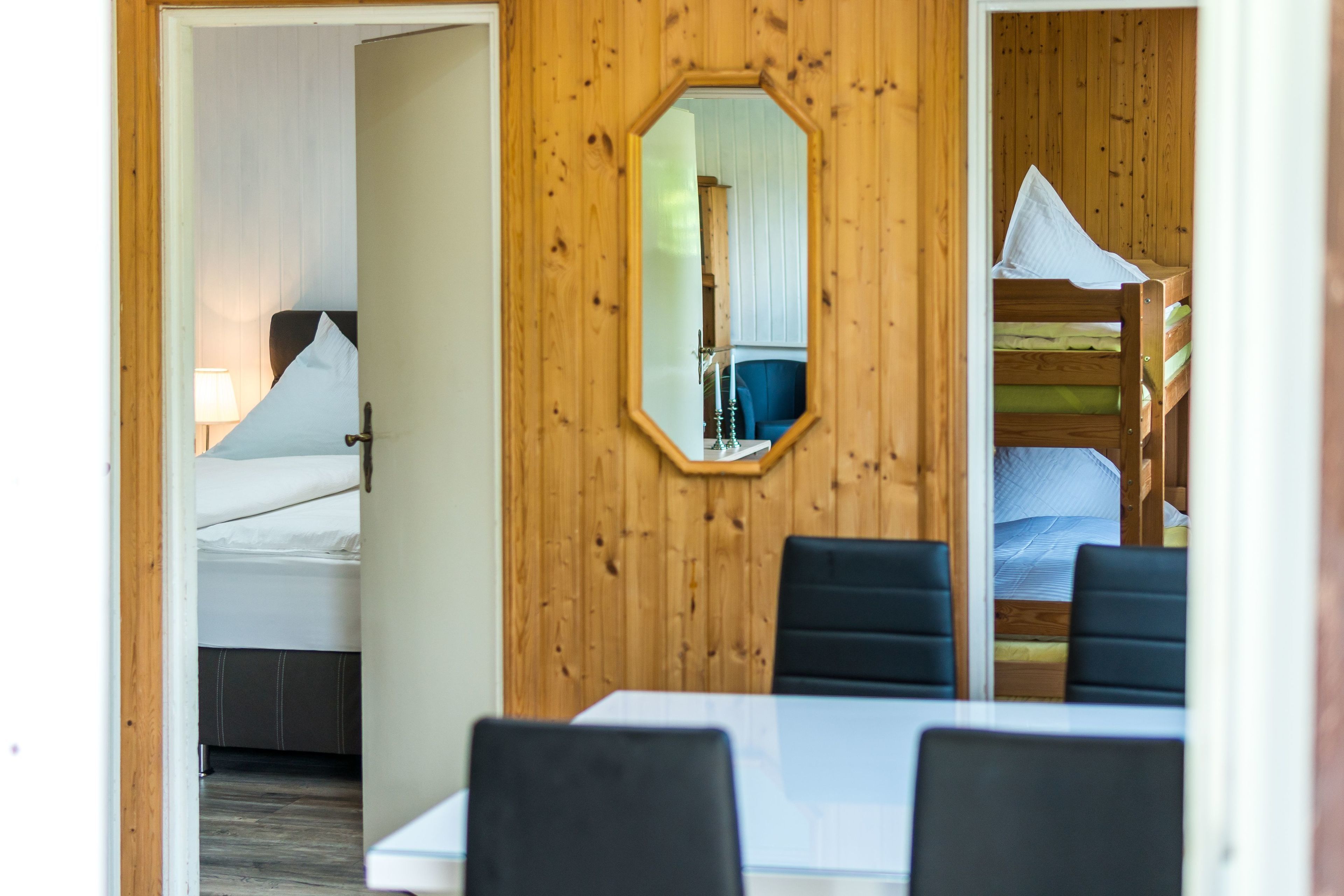 A room with a wooden wall, a bed with white bedding, a table with chairs, and a mirror.