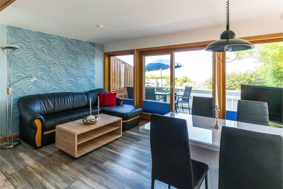 A living room with a sofa, coffee table, and chairs, a dining table with chairs, a patterned wall, and a view through large windows.