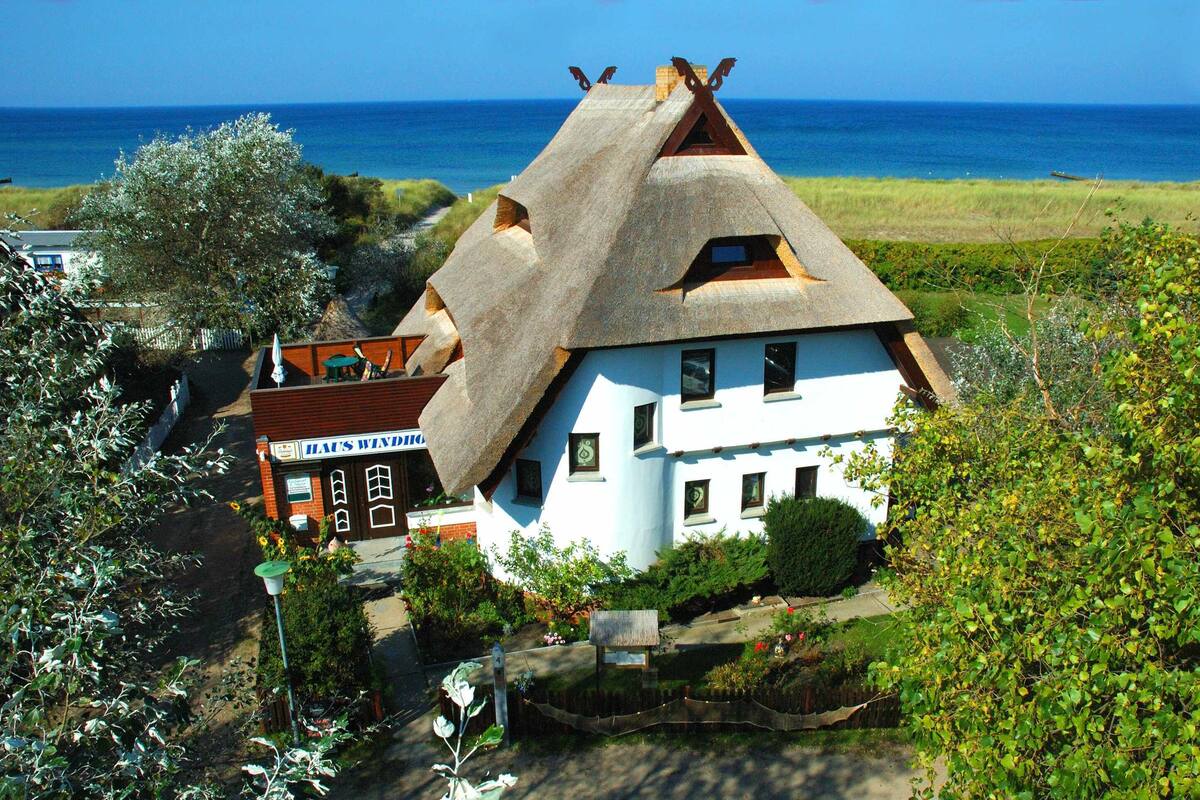 A traditional thatched-roof house with a white facade, a visible garden, and a view of the sea.
