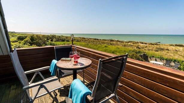 Terraza de madera con una mesa redonda y dos sillas, con vistas a una zona verde y al mar.