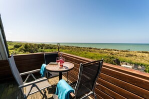 Terraza de madera con una mesa redonda y dos sillas, con vistas a una zona verde y al mar.