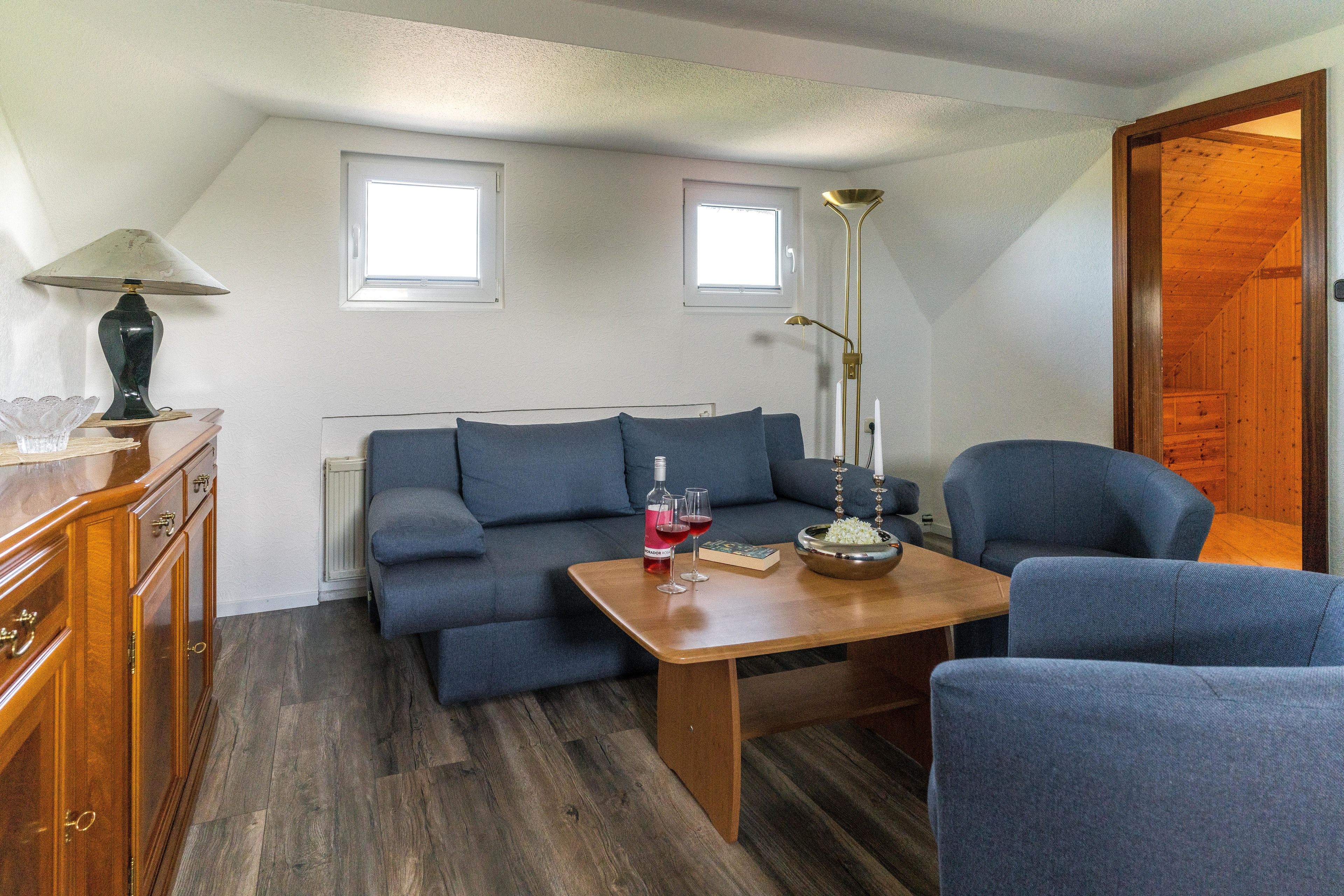 A cozy living room with a sofa, a wooden table, and a lamp. There are two windows providing natural light.