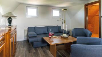 A cozy living room with a sofa, a wooden table, and a lamp. There are two windows providing natural light.
