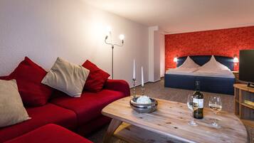 A hotel room with a red sofa, a wooden coffee table with a bottle and glasses, a bed with a red headboard, and a wall with a red floral pattern.
