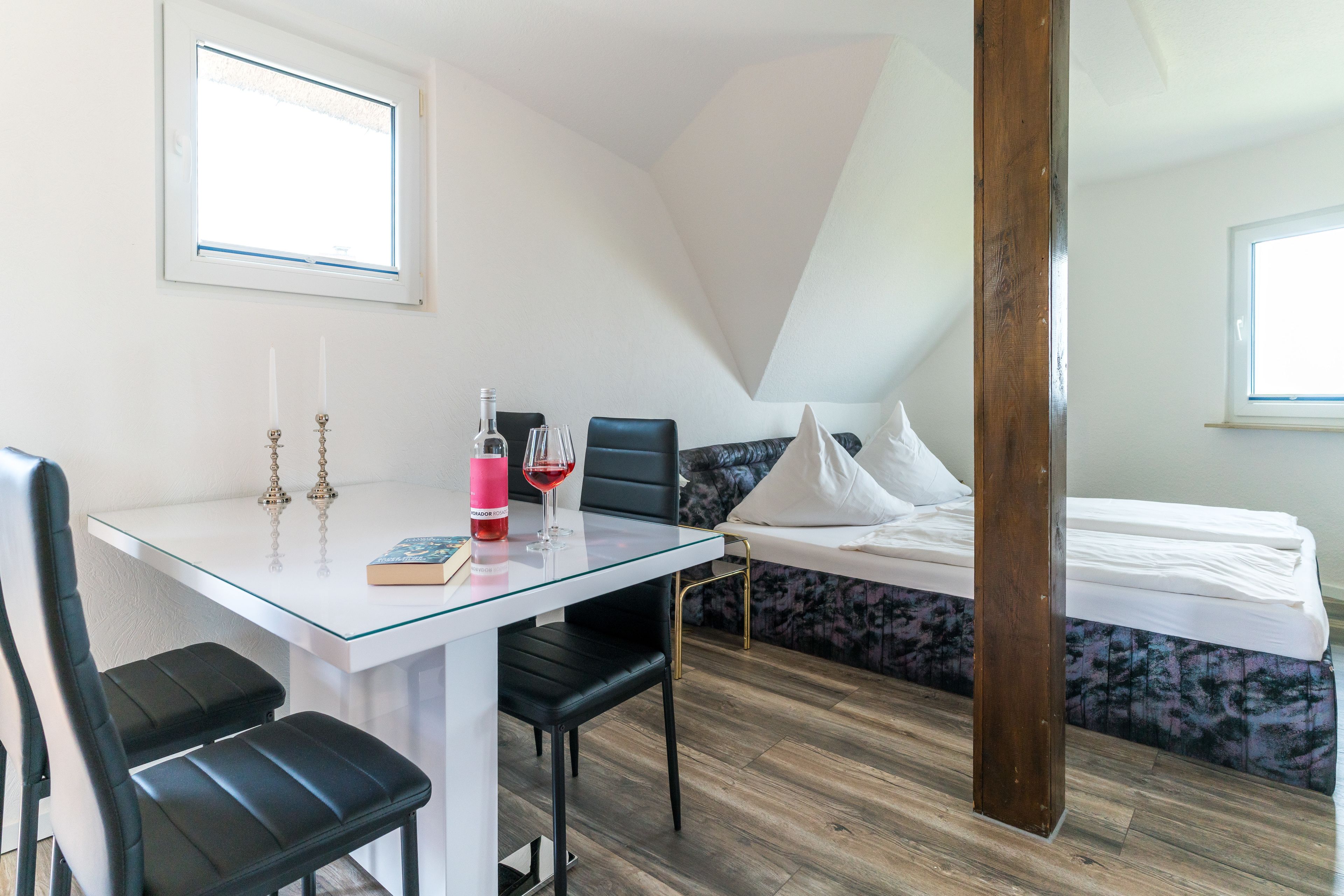 Ein modernes Zimmer mit einem Glastisch, zwei schwarzen Stühlen, einem Bett mit blumigem Überwurf und einem Holzbalken.